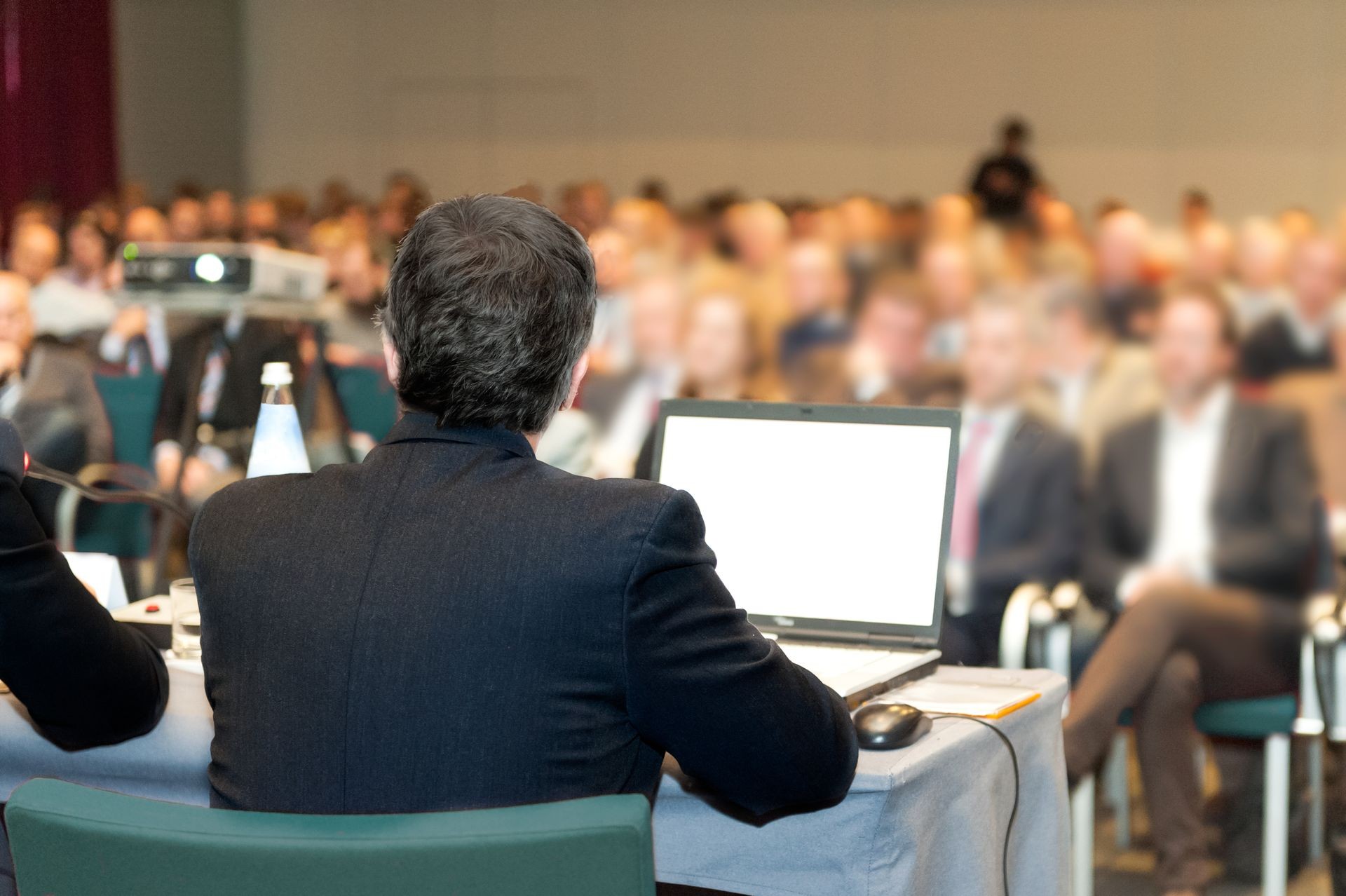 Person sitting at a conference table with a laptop, facing a blurry audience.