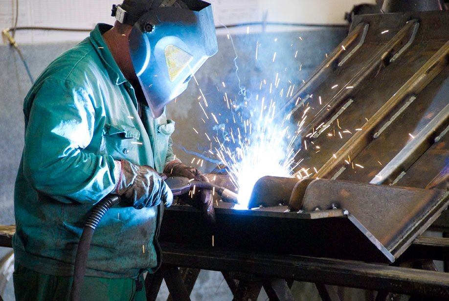 Welder in protective gear working with sparks flying from welding tool on metal surface.