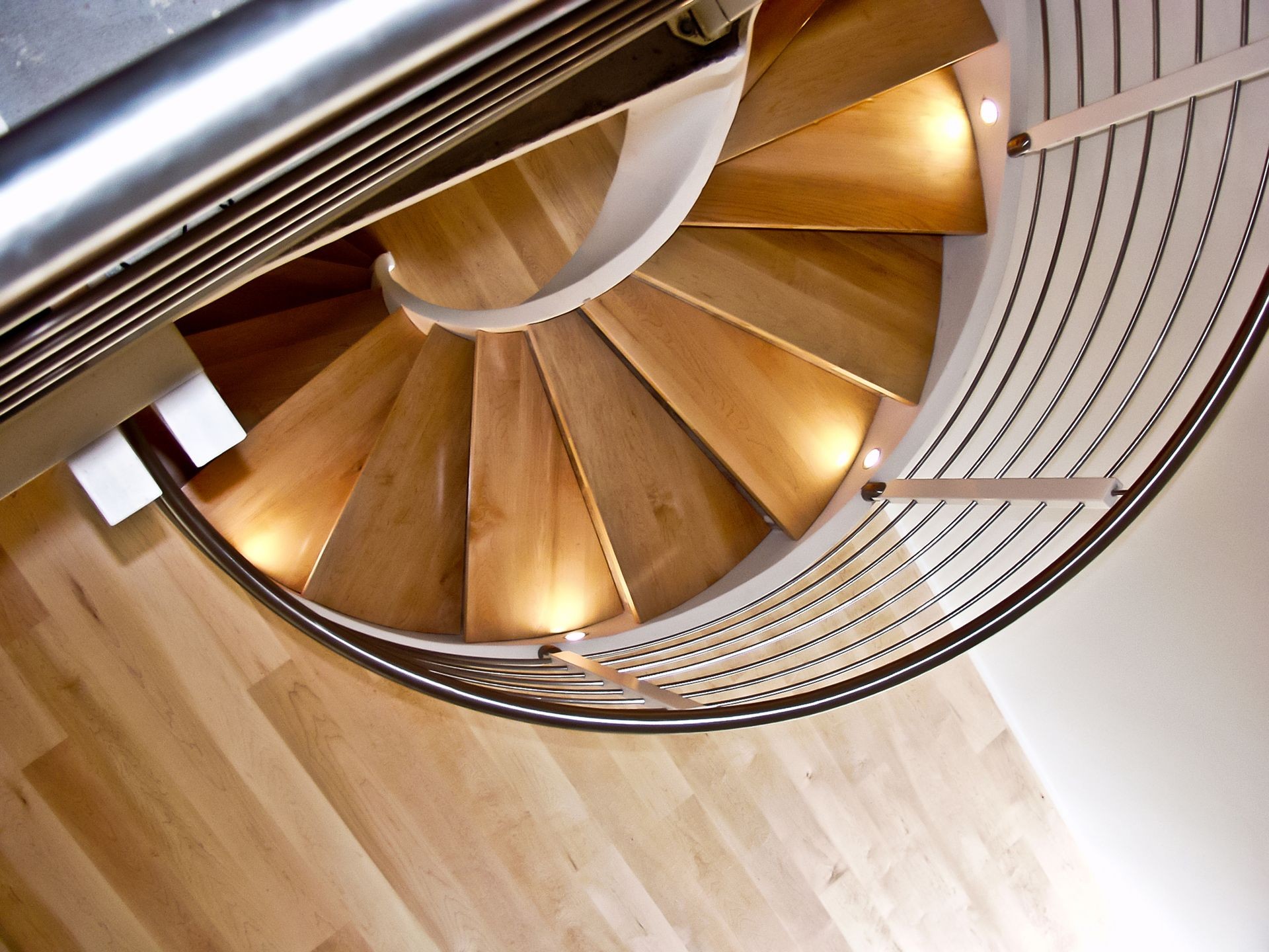 Top-down view of a wooden spiral staircase with metal railings and steps illuminated by recessed lights.